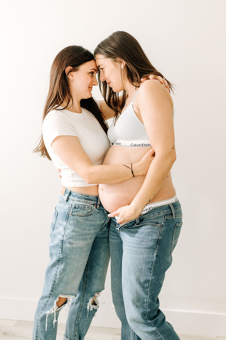 woman in white t shirt and jeans hugs her pregnant wife as their foreheads touch