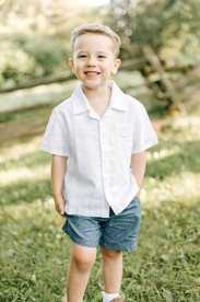 A young boy in a short sleeve white button down shirt and blue shorts stands in a grass field and smiles