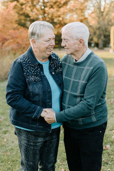 An older couple hug and look at each other smiling on a grassy path under trees in the fall