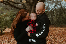 A man and woman look at the baby in the man's arms while they stand under an autumn tree