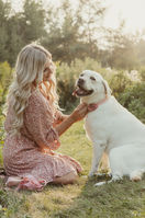 A woman in a pink dress sits on grass and pets a labrador retreiver