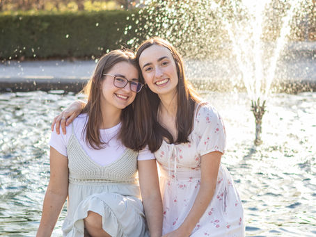 Two woman sit in front of a fountain holding hands