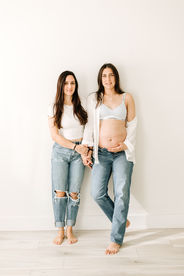 two woman hold hands as they lean against a plain wall, one woman in white t shirt and jeans and the other in jeans, a white bra and white button down shirt and pregnant stomach