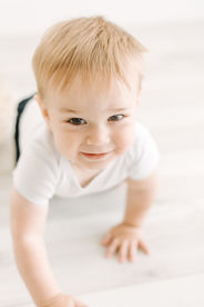 A one year old boy in a white onesie and dark pants crawls on the floor
