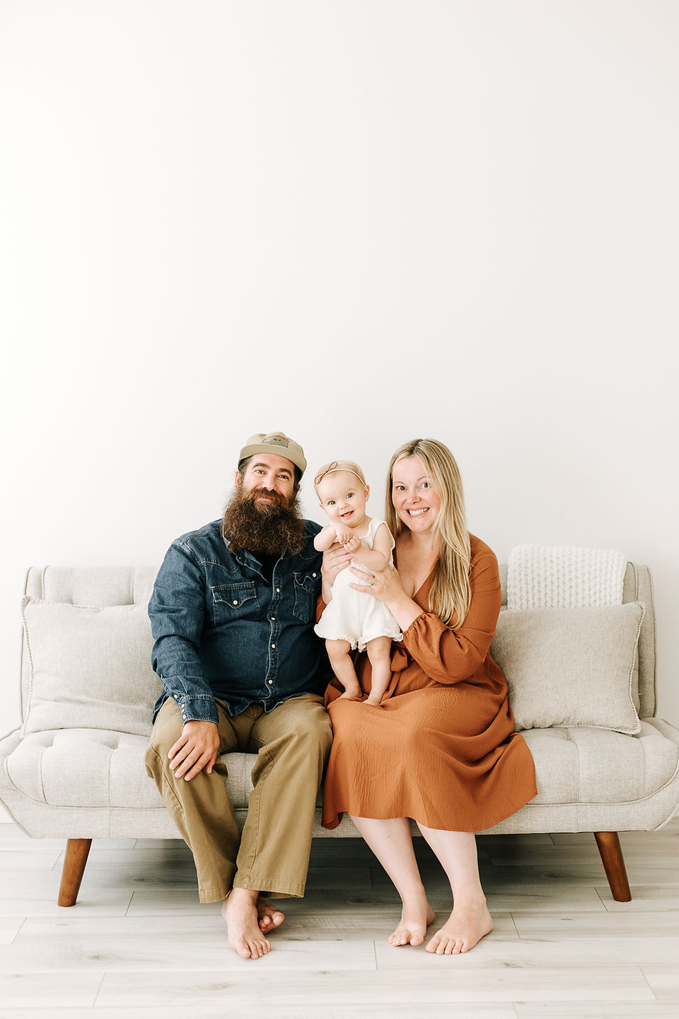 A man and woman sits on a couch and hold a one year old girl standing on their lap
