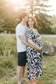 A man wraps his arms around a pregnant woman and kisses her forehead in a green field while the sun sets behind them