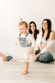 A toddler girl smiles at the camera as she walks, with two woman sit on the floor watching her and smiling
