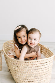A young girl hugs a toddler boy from behind as they sit in a moses basket