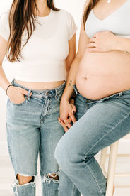 torsos of two woman, one in a white t shirt and jeans with one had in her pocket and the other hand holding her wife's hand as her wife sits on a stool and rests her other hand on her pregnant bare stomach