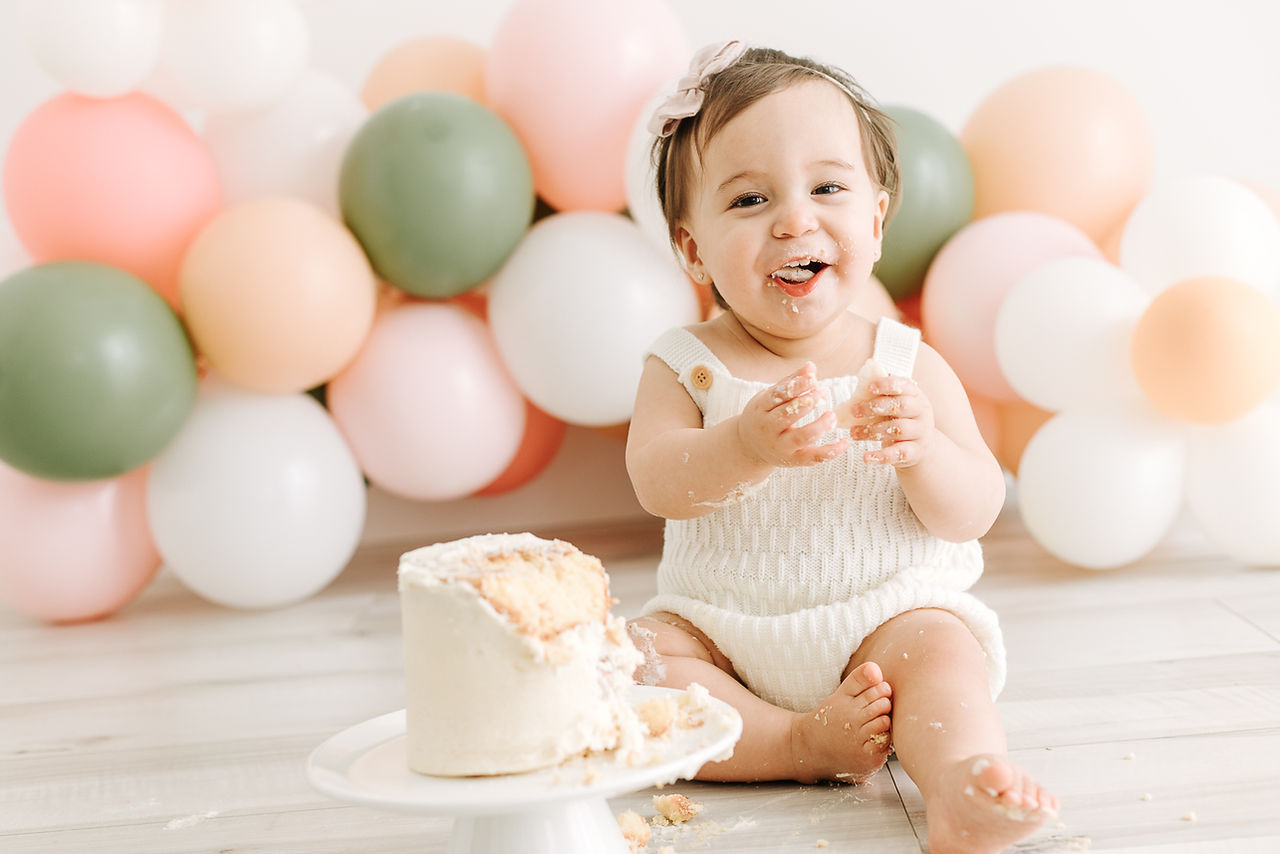 1 year old girl in a white outfit and bow sits in front of balloons and behind a cake on a white dish as she eats some cake