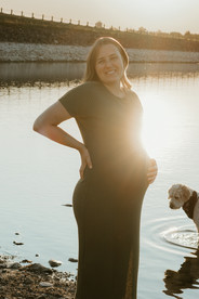 A pregnant woman stands by a lake as her dog swims at sunset