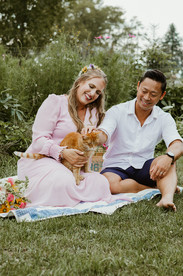 A woman and man sit on a blanket on grass surrounded by flower bouquets and cuddle a kitten