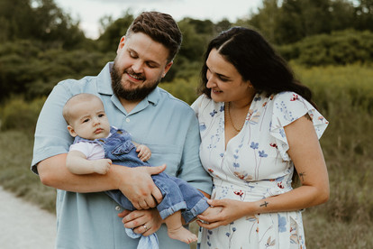 A woman and man stand in a grassy field and look down at the baby in the man's arms