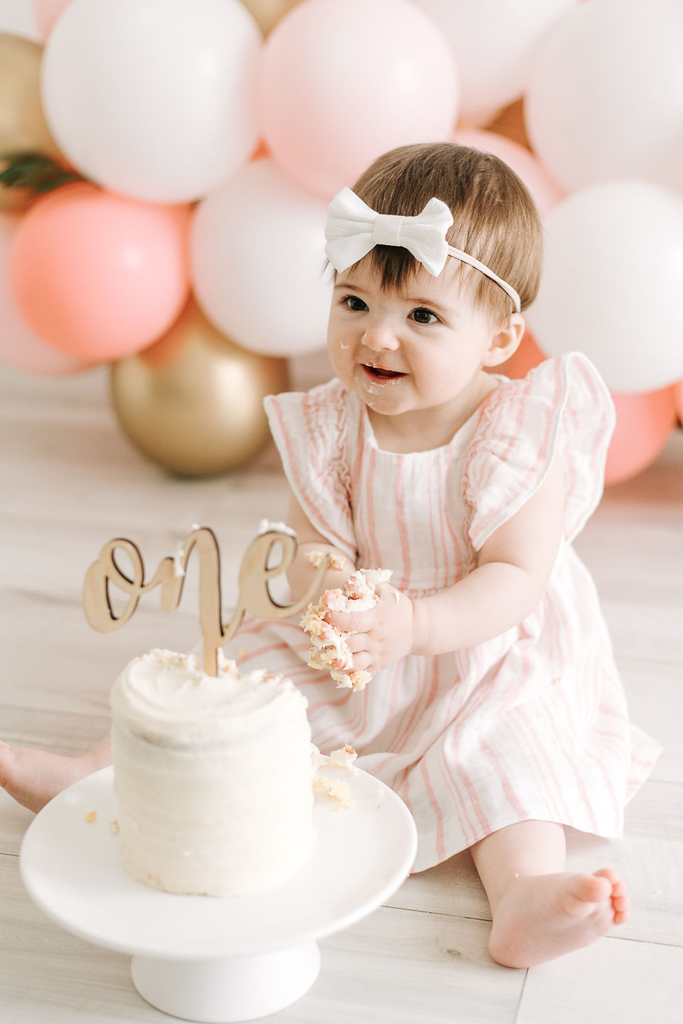 A one year old girl sits on the floor behind a white cake with a cake topper that says one with a balloon garland in the background