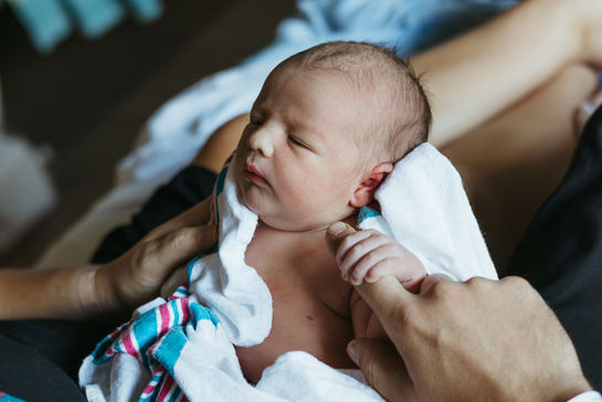 A newborn baby wrapped in a hospital blanket holds onto a man's finger