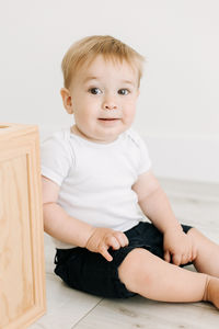 A one year old boy in a white onesie sits beside a large wooden block