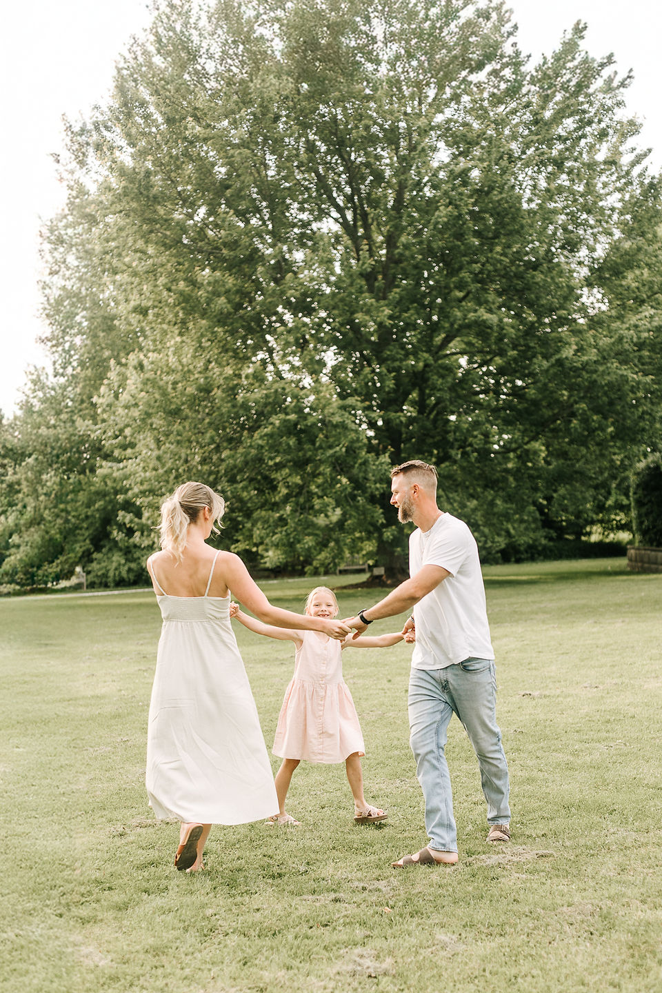 A man, woman, and young girl hold hands in a circle on a grassy field in front of a tree