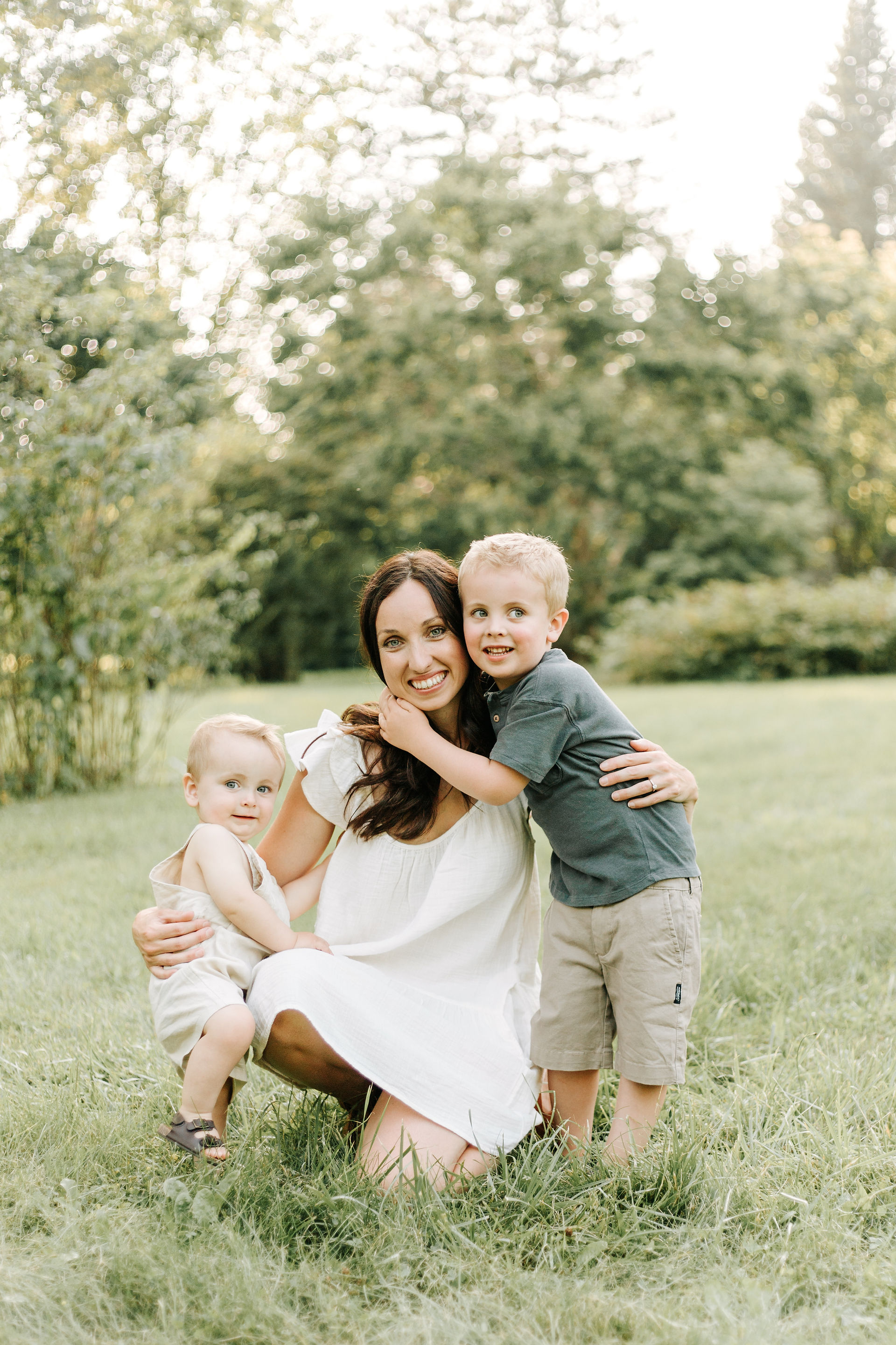A woman in a white dress crouches down and hugs two boys