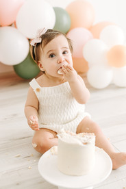A one year old girl sits in front of balloons and eats some cake