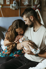 A man sits on the edge of a hospital bed cradling a newborn baby while a woman sits next to him and kisses the baby's head