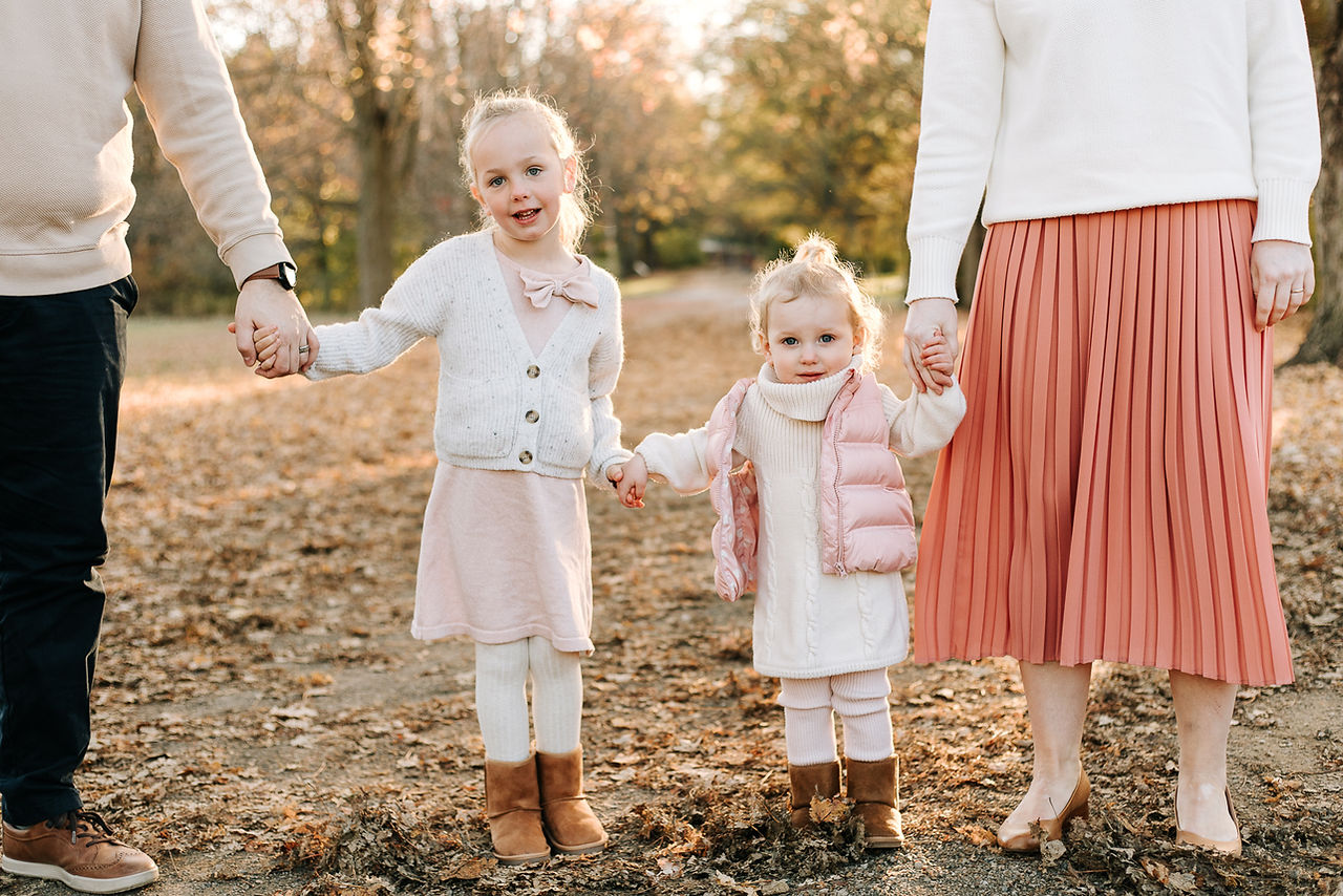 A man and woman stand hand in hand with two young girls on a path in the forest in fall