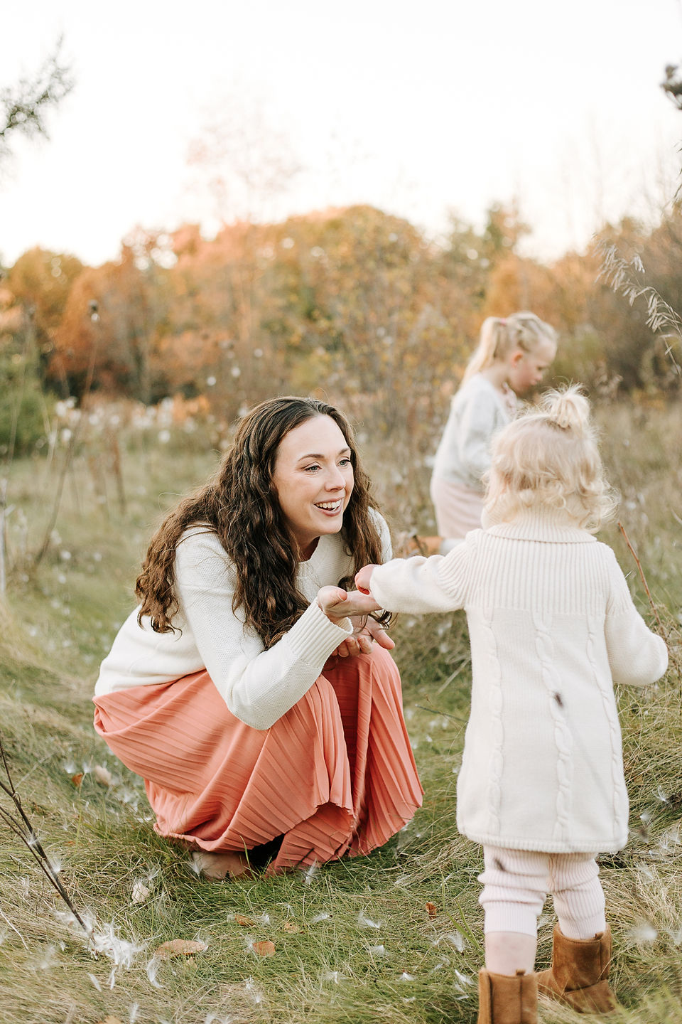 A woman crouches down and smiles at a young girl as they stand in a grass field with milkweed