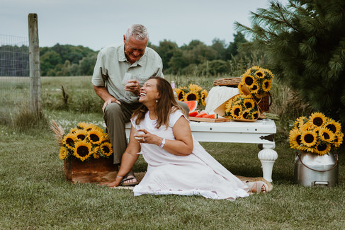 A man sits on a table and clinks wine glasses with a woman sitting at his feet as they smile at each other in a grass field