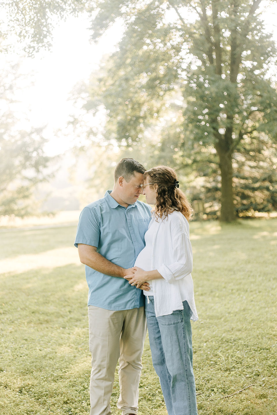A man and pregnant woman rest their foreheads together and rest their hands on her pregnant stomach as they stand in a grass field under trees