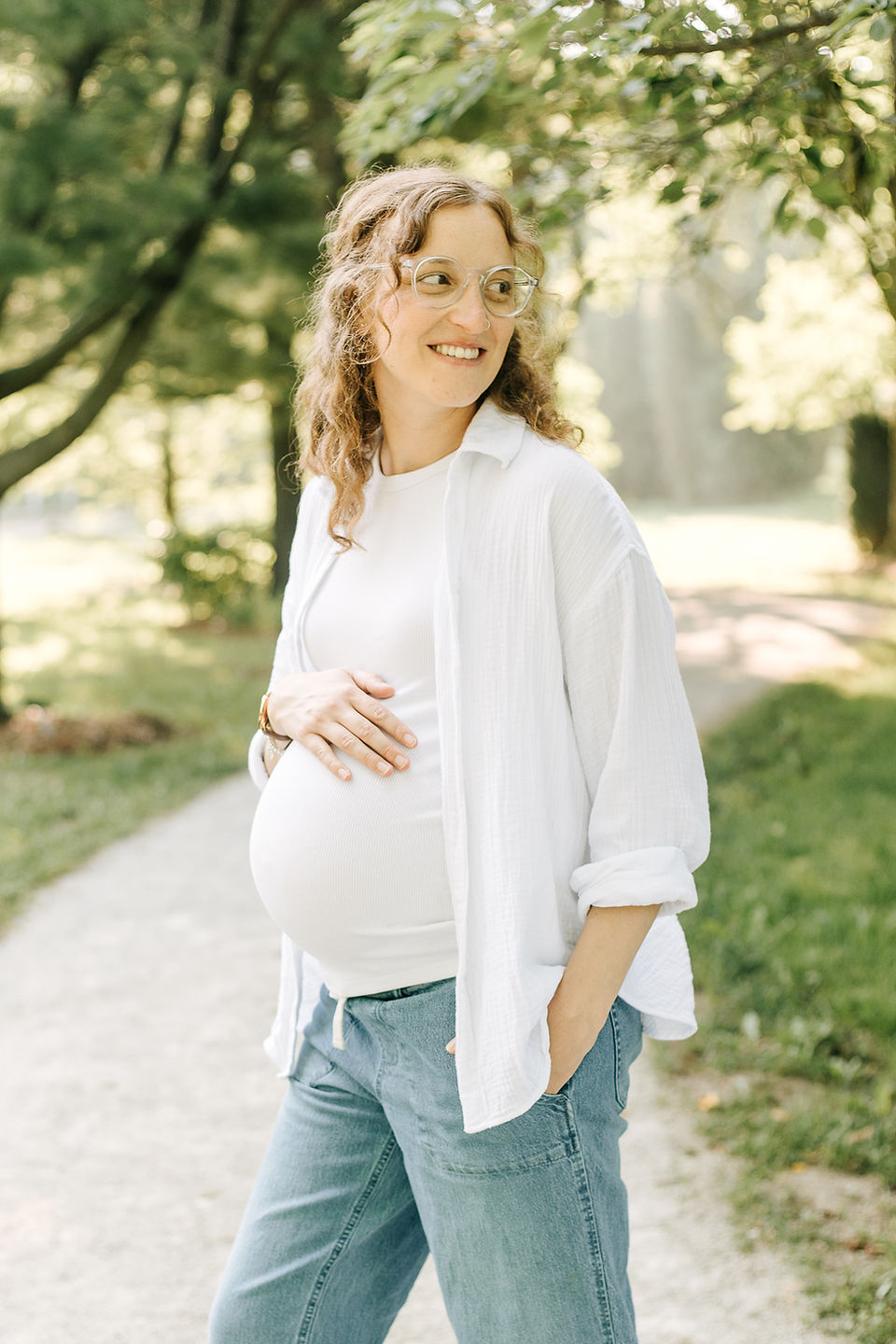 A pregnant woman in jeans and a white shirt rests her hand on her stomach and looks into the distance and stands on a path under trees