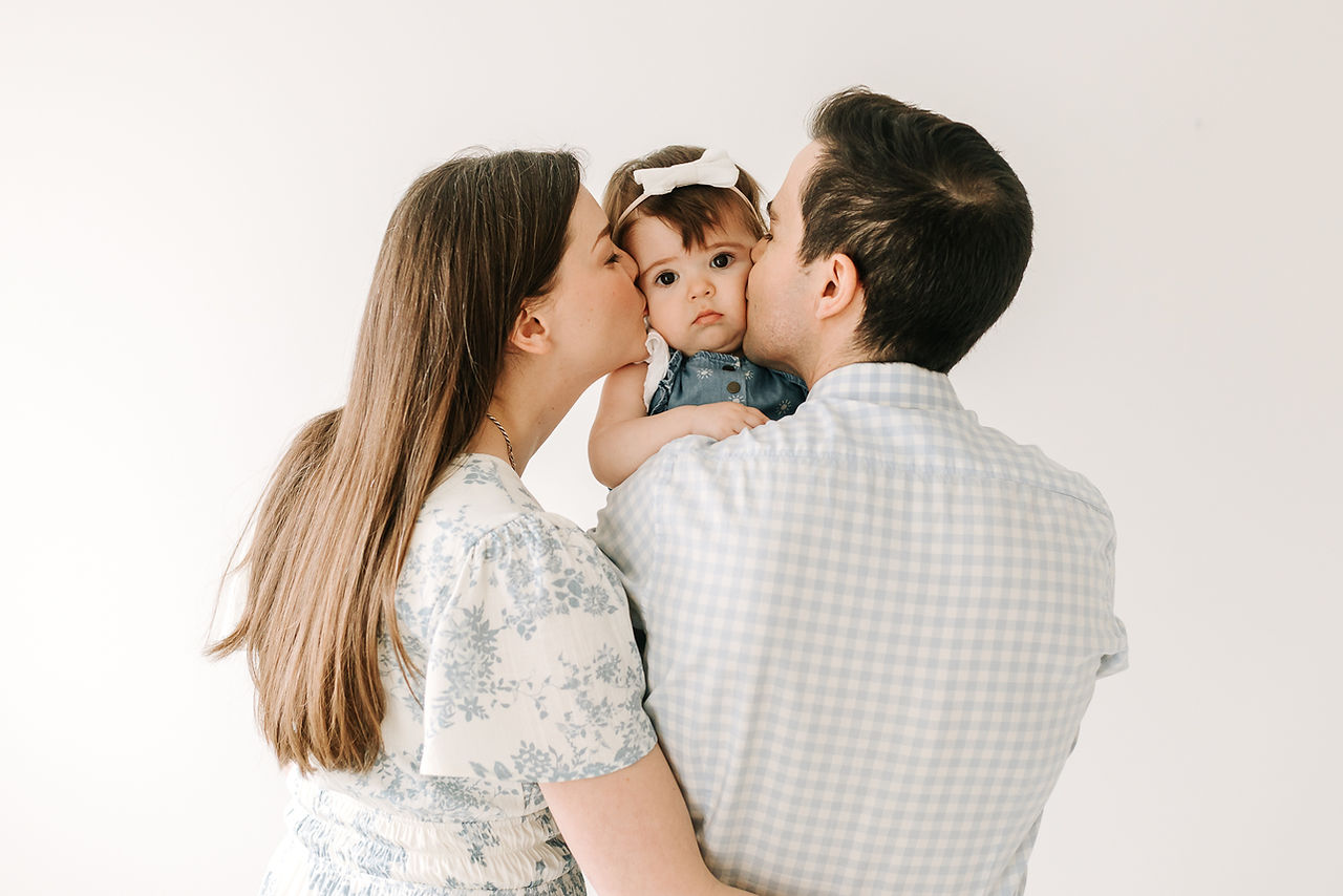 The backs of a woman and man stand beside each other and kiss the cheeks of a girl held between them