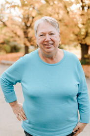 An older woman stands with her hand on her hip and smiles under trees in the fall