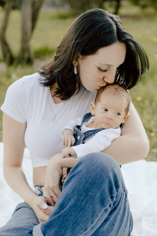 A woman sits on a blanket on grass and kisses the top of a baby's head