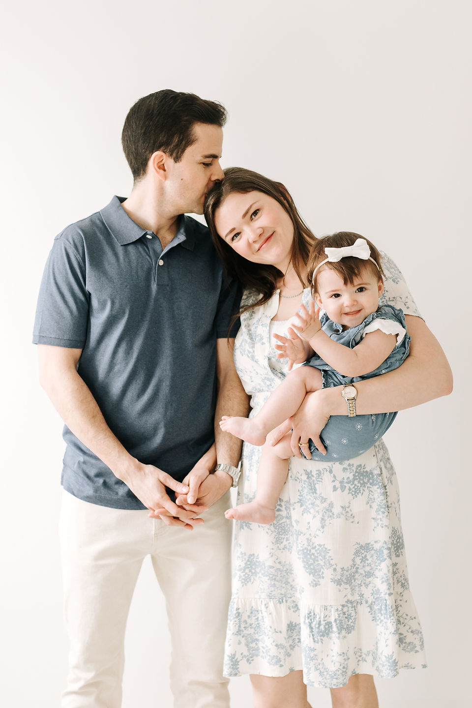 A man kisses the head of a woman who rests her head on his shoulder as she holds a one year old girl in her arms