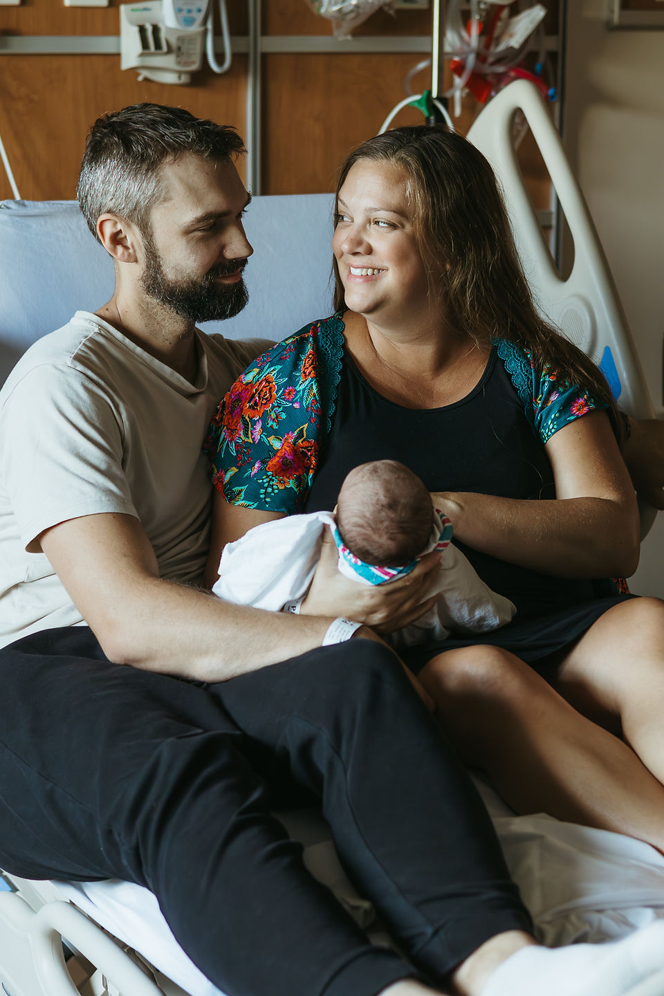 A woman and a man sit on a hospital bed and look at each other as she cradles a newborn baby in her arms