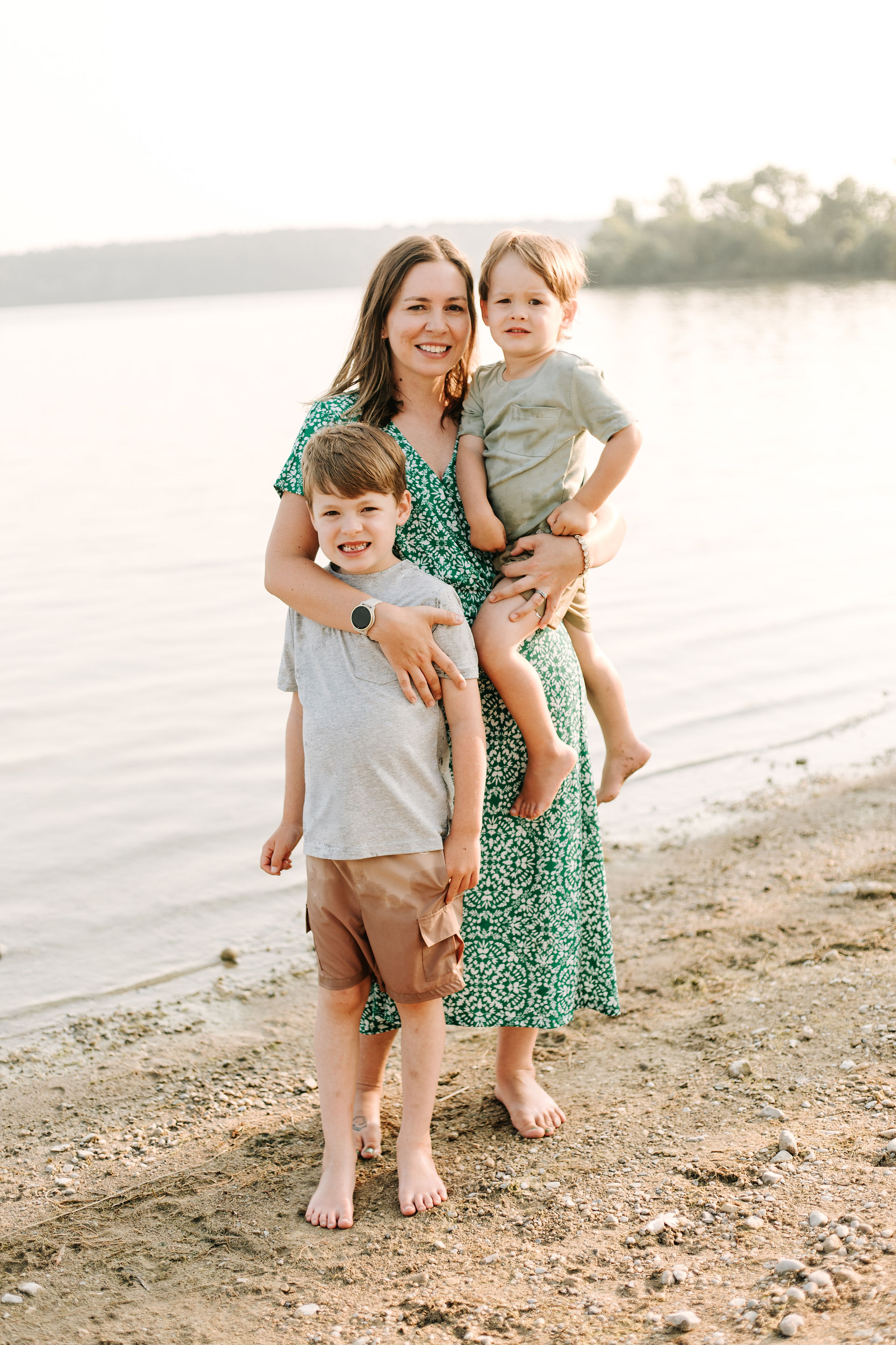 A woman stands on the beach at a lake while she holds a young boy in her arms and hugs another one standing beside her