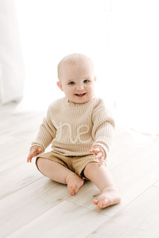 A one year old boy sits on the floor and wears a knit sweater embroidered with the word one