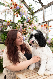 A black and white dog sits on a small wooden bench with a large floral arrangement in the background as a woman kneels next the dog looking at her