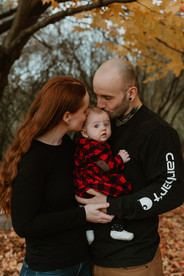 A man and woman stand under a fall tree and kiss the top of a baby's head