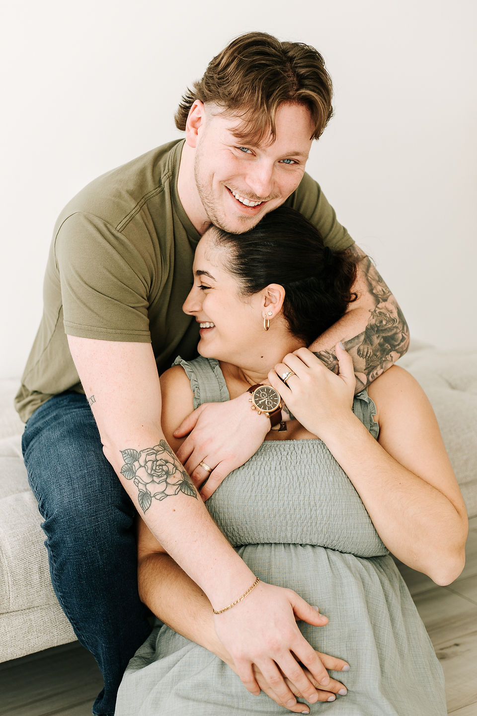laughing pregnant woman sits between her husband's legs as he leans over and hugs her smiling