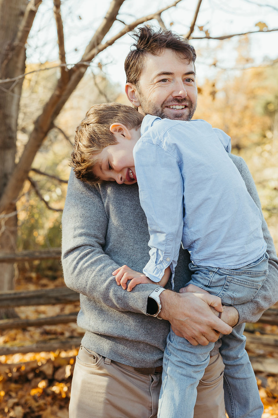 A man holds a boy in his arms as he stands under an autumn tree