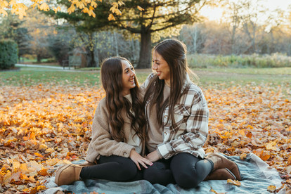 Two teenage girls sit on a blanket in a park in autumn and look at each other and smile