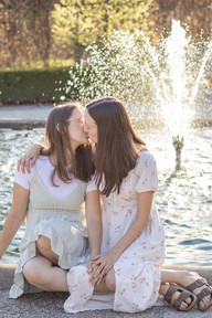 Two woman sit in front of a fountain and hold hand and kiss