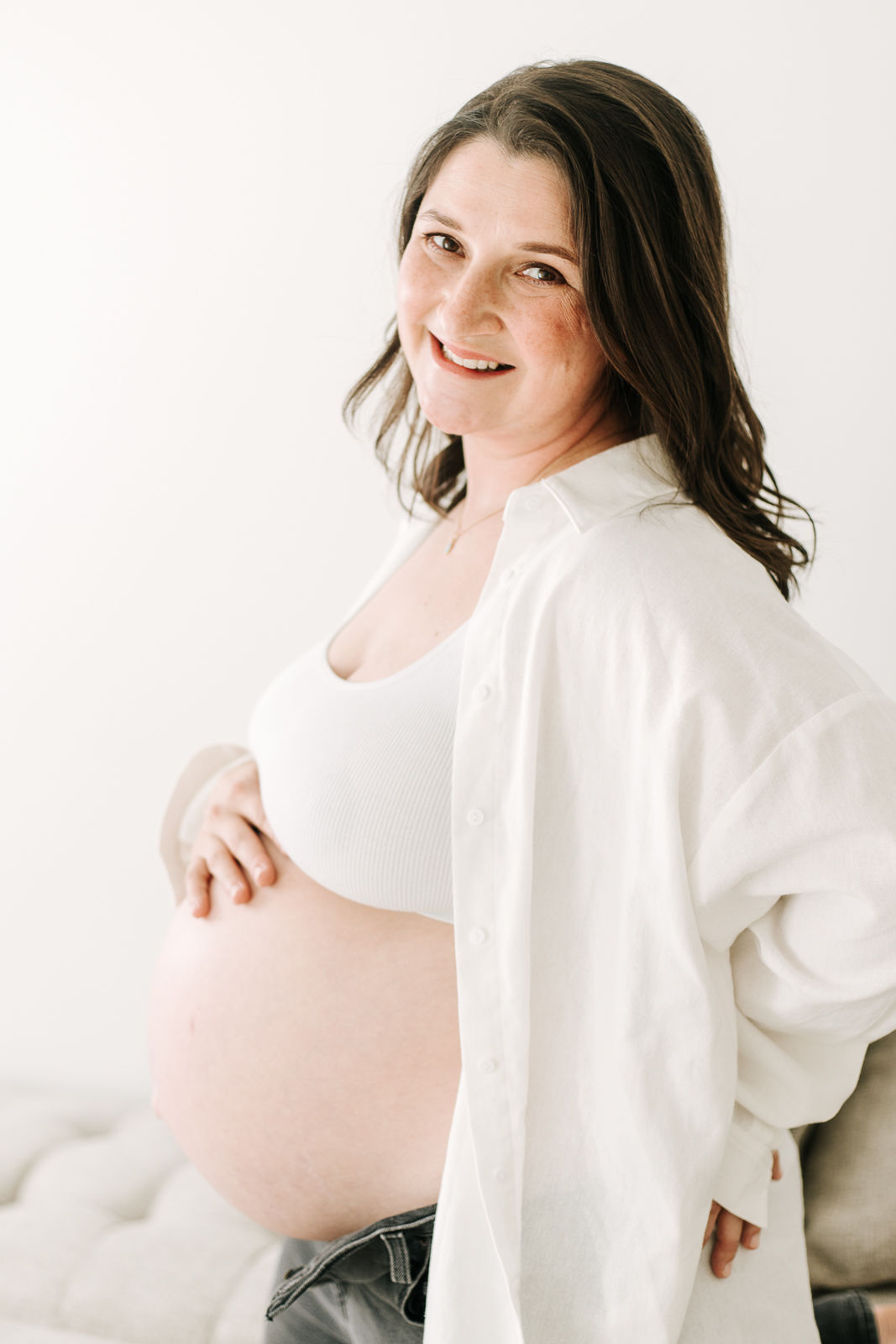 A pregnant woman in a white button down shirt and white bra kneels on a couch with one hand resting on her stomach