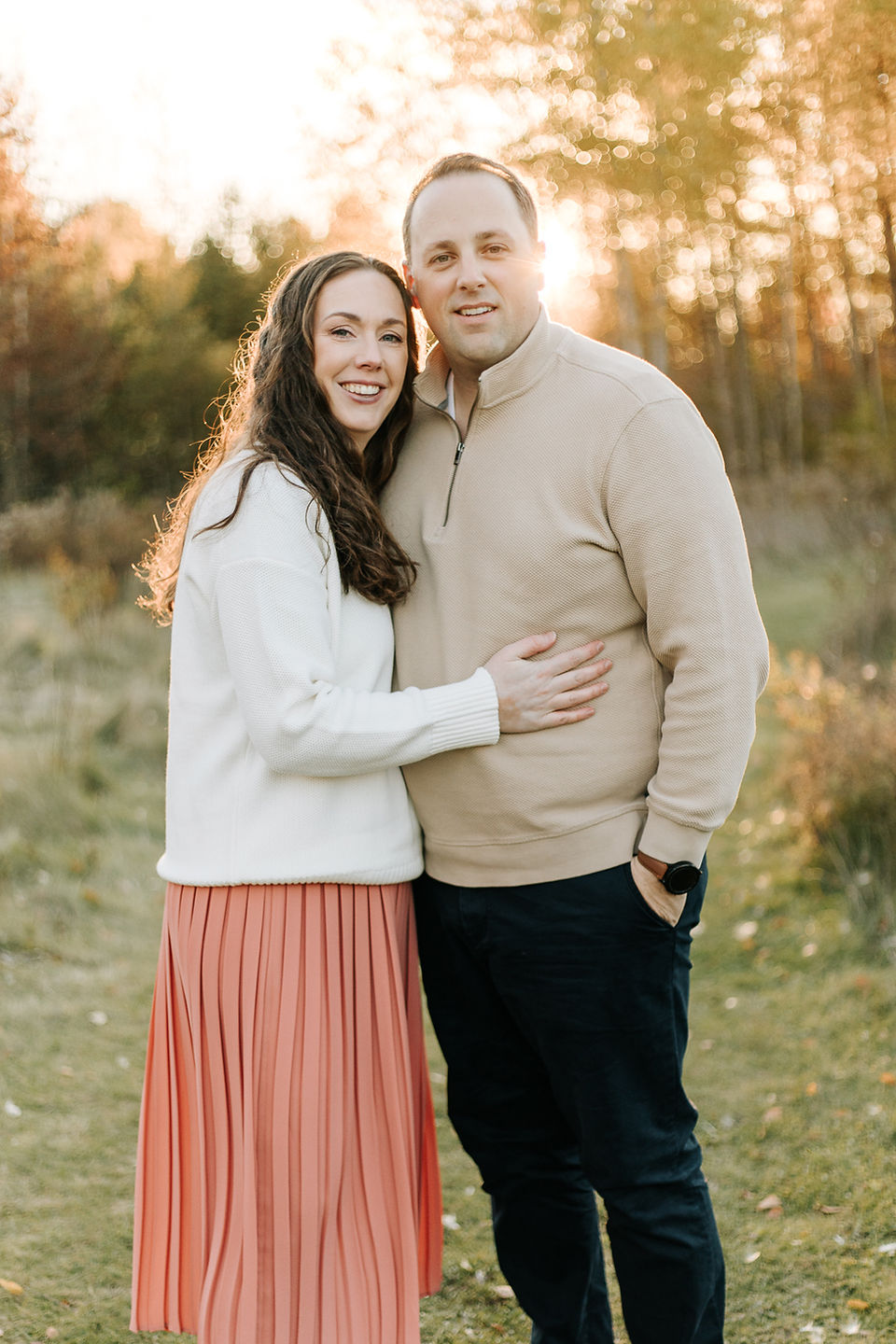 A man and woman hug and smile as they stand on a grass path by trees as the sun sets