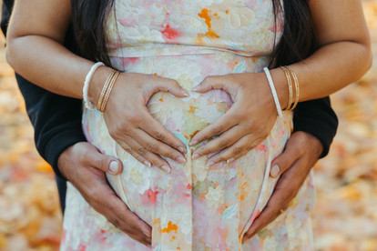 A pregnant woman's stomach as she holds her hands to make a heart shape on it and a man's hands rest underneath her stomach