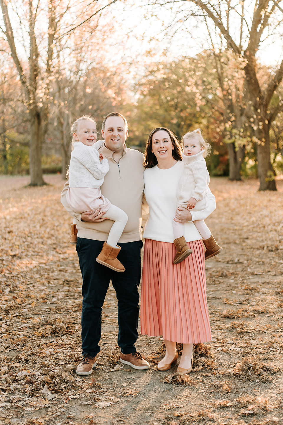 A man and woman each hold a little girl in their arms as they smile on a path under trees in the fall