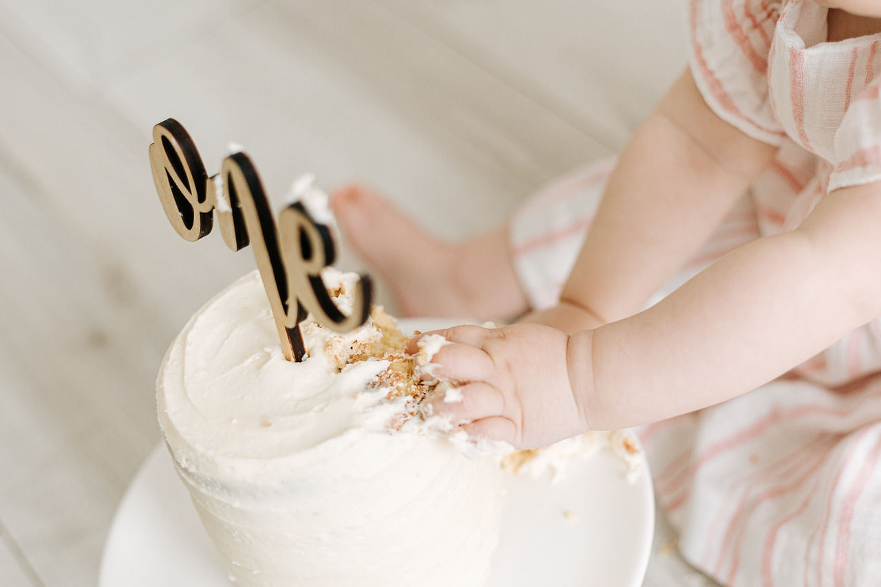 overhead picture of a white cake with a cake topper that says one with one year old hands in the cake