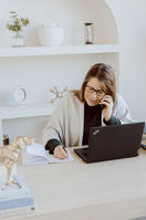 A woman works at a desk as she writes on paper with her laptop open and talking on a cell phone