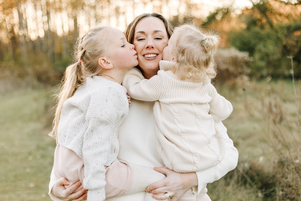 A woman stands with two girls in her arms as they each kiss her cheeks