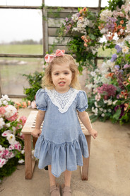 A girl in a blue dress with a pink bow in her hair leans against a small wooden bench in front a large floral arrangement